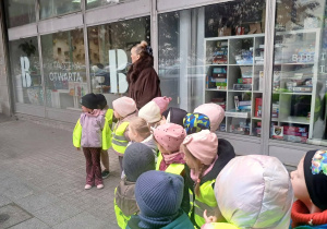 Dzieci z opiekunem, stoją przed budynkiem biblioteki miejskiej. Mają na sobie żółte kamizelki bezpieczeństwa.