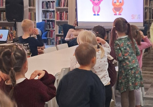 Dzieci stoją przy stoliku i pokazują ruchem części ciała , śpiewane w języku angielskim. Naśladują ruchy postaci wyświetlanych na ekranie multimedialnym.