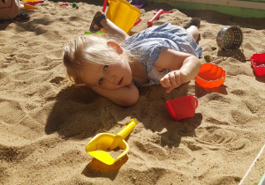 Dziewczynka opala sie w piaskownicy leżąc na piasku.
