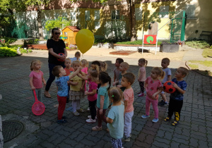Dzieci stoją całągrupą w ogrodzie. Dwoje dzieci trzyma żółty balon wpełniony helem.
