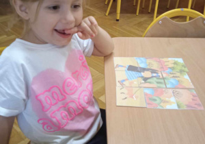 Dziewczynka trzymająca palec w buzi siedzi przy stoliku, na którym znajduje się ułożony przez nią obrazek
