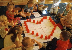 Dzieci siedzą dookoła maty do kodowania na której znajduje sie ułożone z kubków serce