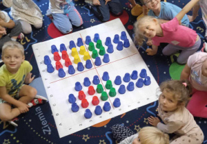 Dzieci siedzą wokół maty do kodowania, na której ułożone są sygnalizatory świetlne z kolorowych kubków