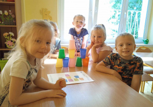 Dzieci siedzą przy stoliku na którym znajduje się wieża z kolorowych kubków
