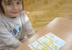Uśmiechnięta dziewczynka siedzi przy stoliku na którym leży ułożony przez nią obrazek przedstawiający mapę Europy