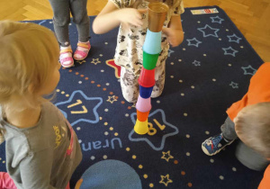 Dziewczynka układa na dywanie wieżę kolorowych kubeczków