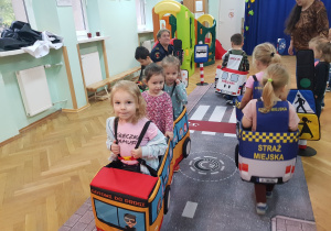 Przedszkolaki jeżdżą w zabawowych pojazdach po makiecie ulicy.
