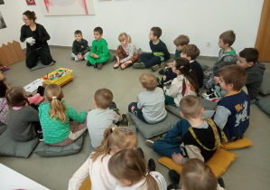 Dzieci siedzą na poduszkach i słuchają pani przewodnik o zasadach panujących w galerii.