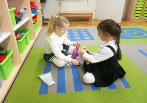 Dziewczynki siedzą na dywanie i zgodnie bawią się lalkami.