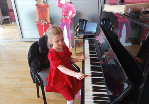 Dziewczynka w czerwonej sukience gra na pianinie.