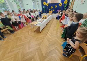 Przedszkolaki w białych bluzkach i białych koszulach oraz w czarnych spódniczkach i czarnych spodniach stoją i recytują wiersze z okazji Dnia Nauczyciela. Na środku sali stoi stół zastawiony jedzeniem.