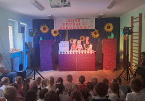 Teatrzyk lalek, czerwona scena, lalki: gospodyni, dziewczynka, kózka. Dzieci na widowni.